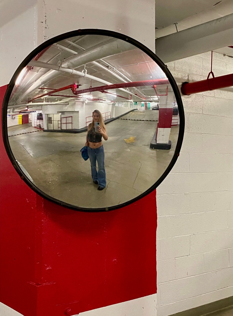 sabrina takes a selfie in front of a convex round mirror in a parking garage, with red and white color blocking on the wall surrounding the mirror. Sabrina wears bell-bottom jeans and a grey crop top. 