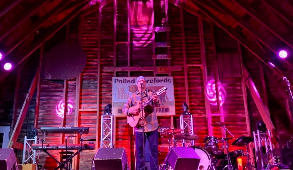 Craig Honeycutt, lead singer of the band Everything, plays guitar and sings into a mic on a stage in a rustic barn. Red and pink swirly lights light up the wooden stage.