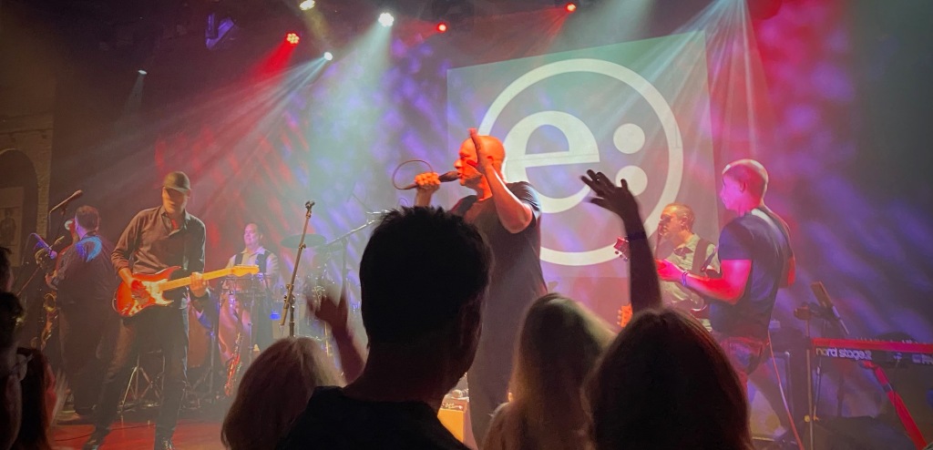 Six or seven members of the band Everything performing on a brightly lit stage, with their logo of a lowercase e: in a circle as a backdrop. members of the audience are in front of the stage dancing.